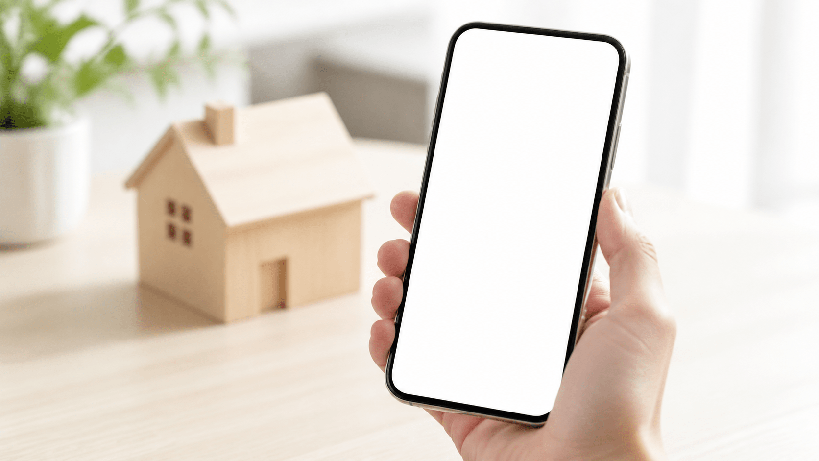 スマートフォンを持つ手と、背景にある家の模型。不動産アプリのイメージ