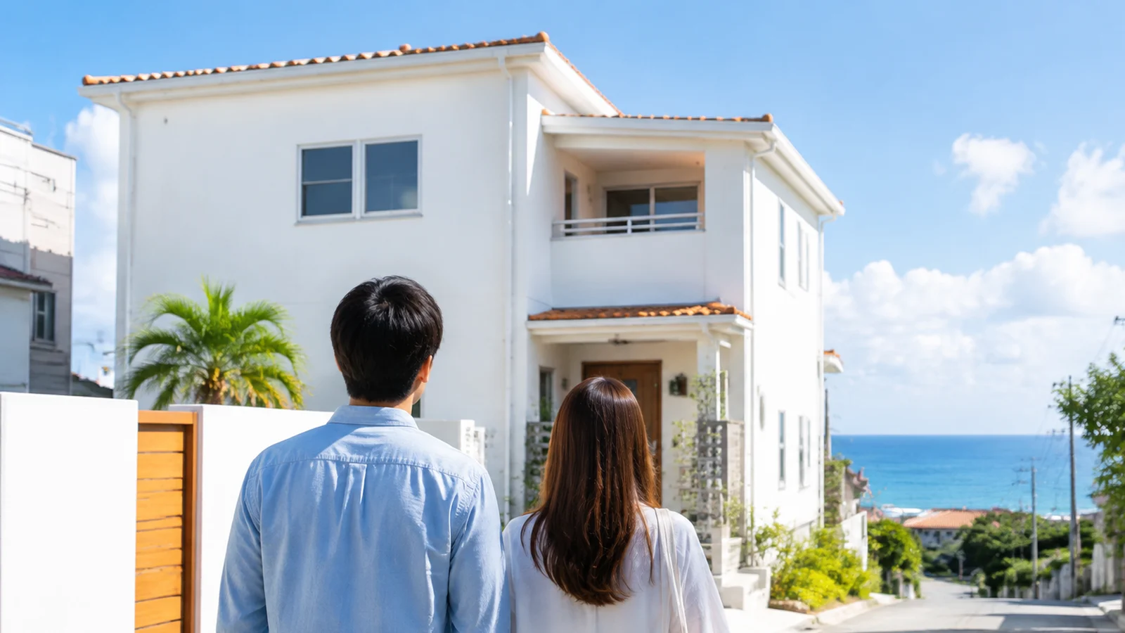 沖縄の住宅街で、新しい白い二階建ての家の前に立つ夫婦の後ろ姿。遠くに青い海