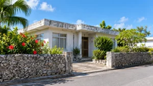 石垣とハイビスカスの前に建つ、年数のたった平屋のコンクリート住宅